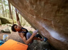 Boulderer making a dynamic move on an outdoor boulder, with crash pad, chalk bucket, and shoes visible, representing the ultimate guide to bouldering gear, grades, and moves.