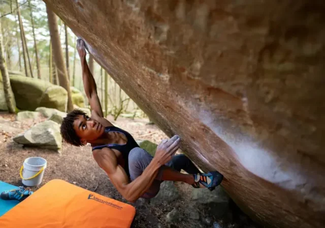Boulderer making a dynamic move on an outdoor boulder, with crash pad, chalk bucket, and shoes visible, representing the ultimate guide to bouldering gear, grades, and moves.