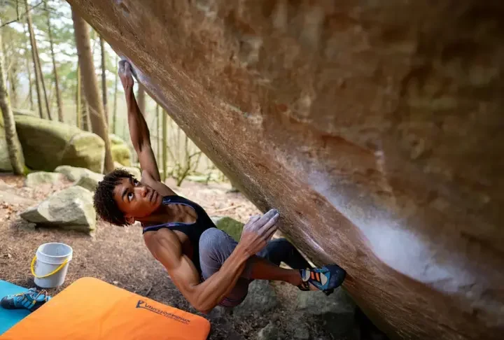 Boulderer making a dynamic move on an outdoor boulder, with crash pad, chalk bucket, and shoes visible, representing the ultimate guide to bouldering gear, grades, and moves.