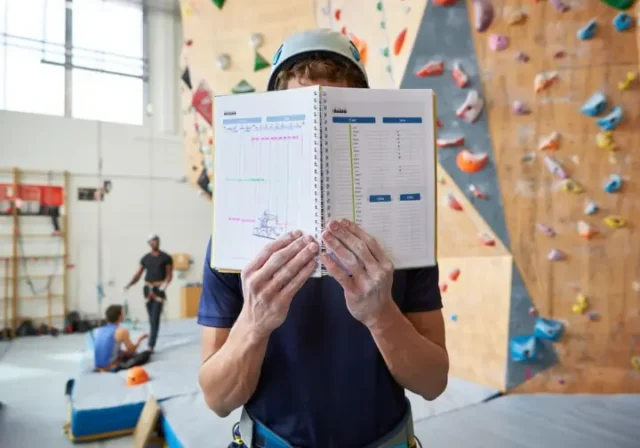 Climber with a notebook planning their session in front of a large indoor climbing wall, illustrating an effective indoor climb plan.