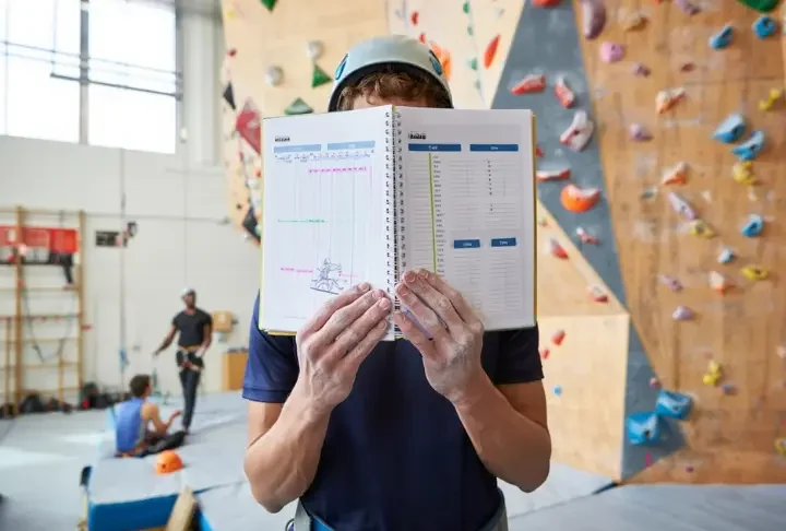 Climber with a notebook planning their session in front of a large indoor climbing wall, illustrating an effective indoor climb plan.