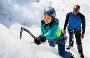 Ice Climbing Kickstart: Gear & Foundational Moves Beginner ice climber making a foundational ice axe swing on an ice wall, guided by an instructor, showcasing essential gear and moves for an ice climbing kickstart.