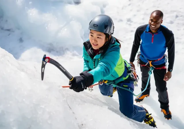 Beginner ice climber making a foundational ice axe swing on an ice wall, guided by an instructor, showcasing essential gear and moves for an ice climbing kickstart.