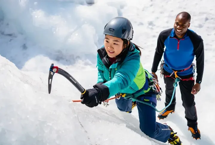 Beginner ice climber making a foundational ice axe swing on an ice wall, guided by an instructor, showcasing essential gear and moves for an ice climbing kickstart.