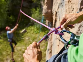 Climber's hands expertly tying a Figure-Eight Follow-Through knot on a harness, with a belayer in the background, mastering essential knots and ropework for sport climbing safety.