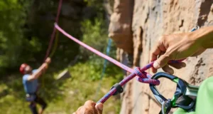 Master Sport Climb Safety: Knots & Ropework Now Climber's hands expertly tying a Figure-Eight Follow-Through knot on a harness, with a belayer in the background, mastering essential knots and ropework for sport climbing safety.