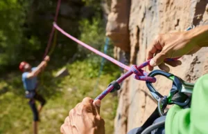 Master Sport Climb Safety: Knots & Ropework Now Climber's hands expertly tying a Figure-Eight Follow-Through knot on a harness, with a belayer in the background, mastering essential knots and ropework for sport climbing safety.
