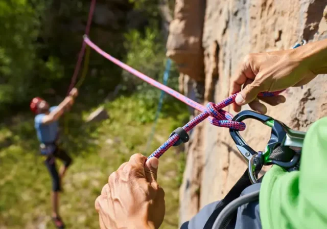 Climber's hands expertly tying a Figure-Eight Follow-Through knot on a harness, with a belayer in the background, mastering essential knots and ropework for sport climbing safety.