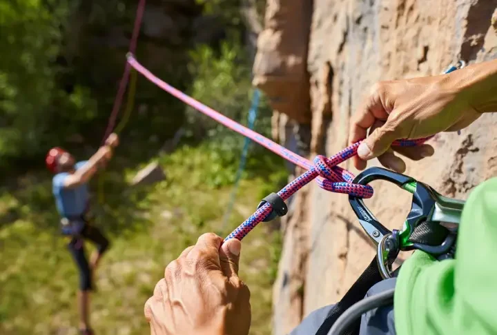 Climber's hands expertly tying a Figure-Eight Follow-Through knot on a harness, with a belayer in the background, mastering essential knots and ropework for sport climbing safety.