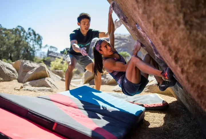 Boulderer having a fall safely guided by an attentive spotter onto well-placed crash pads at an outdoor boulder, illustrating mastering spotting and crash pad usage for safer bouldering.