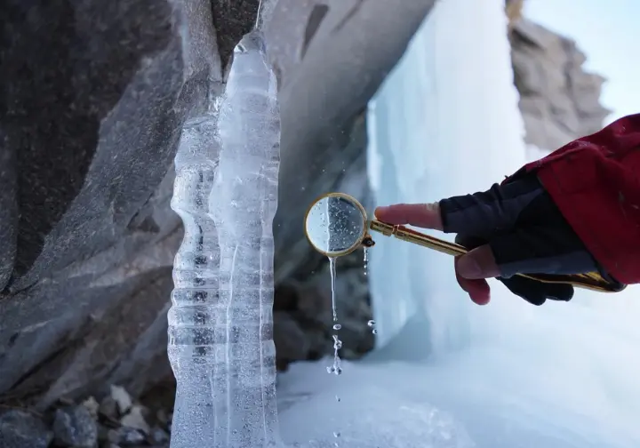 Close-up of ice formation process on a rock face, with a hand examining ice crystals, illustrating the science of ice formations and their properties for climbers.