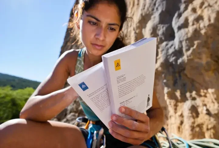 Climber studying a guidebook at an outdoor crag, comparing YDS, French, and UIAA sport climbing grades to understand different global systems.
