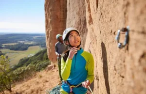 Unlock Success: Read Sport Routes Onsight, Redpoint Climber intently reading a sport climbing route from the ground, planning for onsight or redpoint success, with her belayer in the background at an outdoor crag.