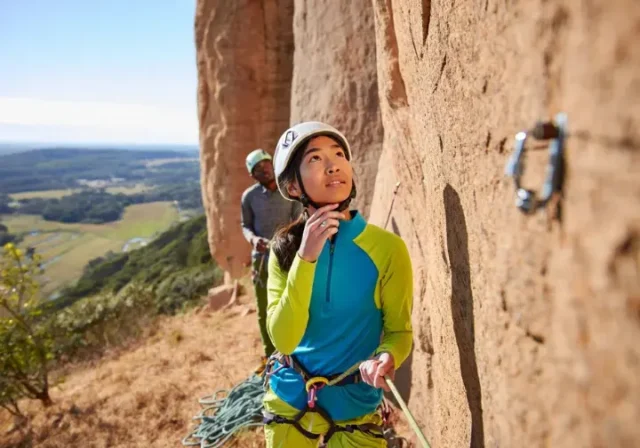 Climber intently reading a sport climbing route from the ground, planning for onsight or redpoint success, with her belayer in the background at an outdoor crag.