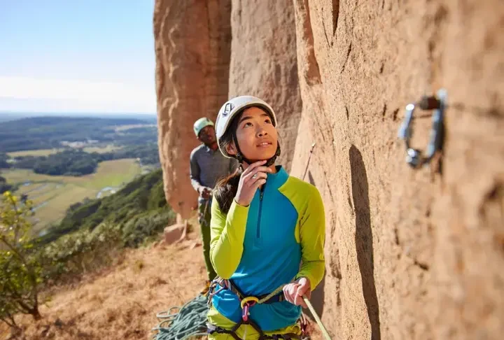 Climber intently reading a sport climbing route from the ground, planning for onsight or redpoint success, with her belayer in the background at an outdoor crag.
