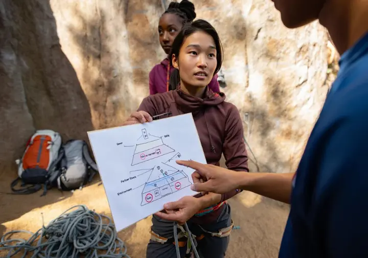 Two diverse climbers at a crag discussing a Swiss Cheese Model diagram, applying it to climbing accidents and safety by identifying layers of defense.