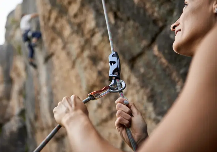 Close-up of a belayer's hands expertly managing the brake strand with an assisted braking device, emphasizing the unseen link of correct belaying to prevent errors.