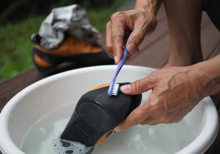Hands gently washing a climbing shoe in a basin, with another shoe stuffed with newspaper air-drying in the shade, showing the deep clean process.