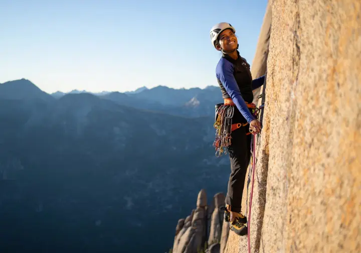 Trad climber with a full rack paused on a granite multi-pitch route, looking out at a mountain vista, embodying the essence of adventure and self-reliance in trad climbing.