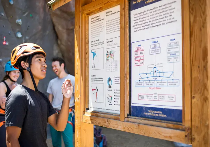 Climber reading about grades, safety, and ethics at an informational kiosk at a sport climbing crag, with other climbers in the background, highlighting essential knowledge.