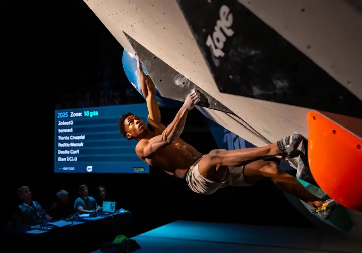 A male climber in an IFSC competition executing a powerful move on a boulder problem, with a 2025 points-based scoreboard in the background, explaining the discipline.