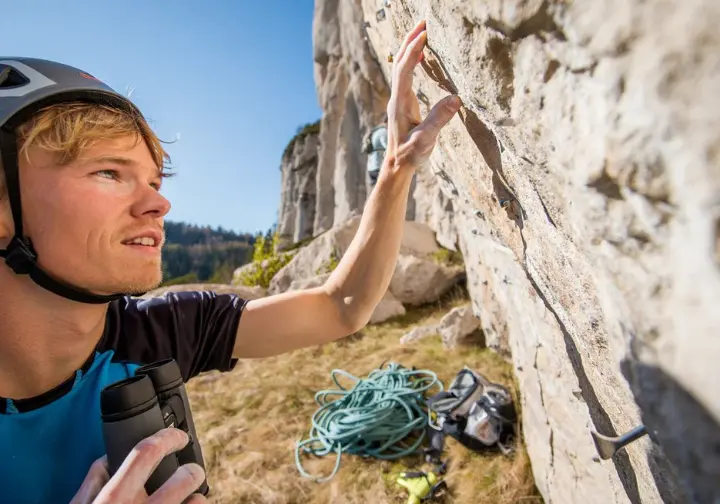 Climber using binoculars to meticulously assess a sport route from the ground, planning strategies for a first-try onsight success at an outdoor crag.
