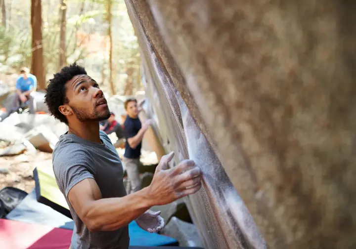 Climber thoughtfully assessing a short boulder problem outdoors, with a crash pad below, illustrating the core concepts of bouldering as rope-free climbing and problem-solving.