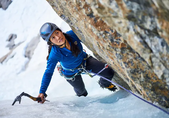 Mixed climber skillfully executing a transition from ice to rock, with one ice tool on ice and the other reaching for a rock hold, demonstrating the art of moving between mediums.