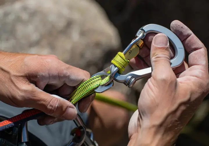 Close-up of hands performing maintenance on a climbing quickdraw, checking both the metal carabiner gate and the textile dogbone sling.