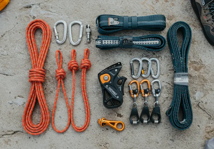 A flat lay of essential climbing gear for improvised partner rescue, including cordelette, Prusiks, locking carabiners, and slings, on a rock surface.