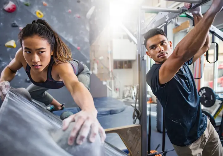 Two diverse climbers in an indoor gym, one practicing footwork on a wall and the other using a hangboard, building their foundational climbing skills.