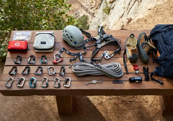 Flat lay of essential outdoor sport climbing kit including rope, harness, helmet, quickdraws, belay device, PAS, shoes, and chalk bag, at the base of a crag.