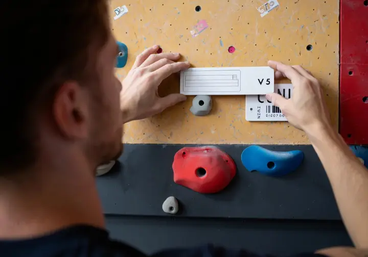 Route setter attaching a grade tag to a new climb, with various hold types nearby, illustrating the language of difficulty and key factors in gym grading systems.