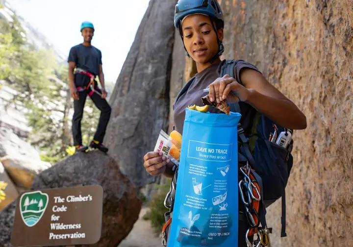 Climber meticulously packing out all trash, including fruit peels, into a reusable bag at a wilderness crag, demonstrating Leave No Trace principles for wilderness preservation.