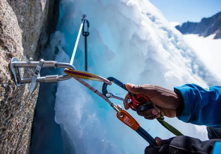 Close-up of a well-built, equalized trad anchor using cams, nuts, and an ice screw on mixed alpine terrain, with a climber's hands checking the master point for alpine security.