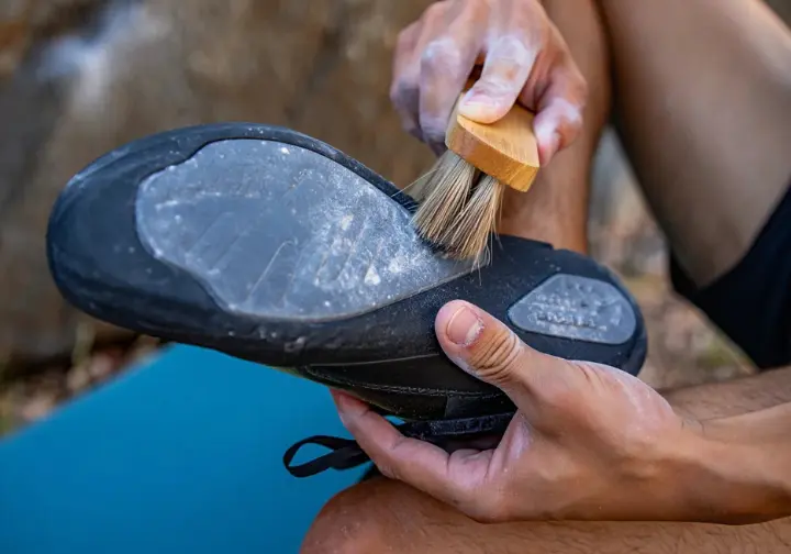 Close-up of a climber using a brush to clean the rubber sole of a climbing shoe to maximize grip and on-wall performance.