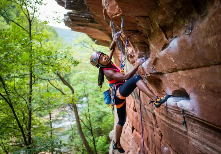 Climber on a steep, pocketed sandstone sport route in a lush forest setting, representing the sandstone paradises of Eastern/Southern US sport climbing crags.