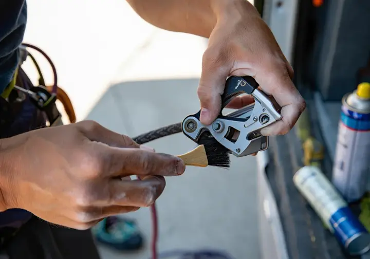 Close-up of a climber performing belay device maintenance, using a small brush to clean the internal mechanism to ensure smooth and safe rope control.