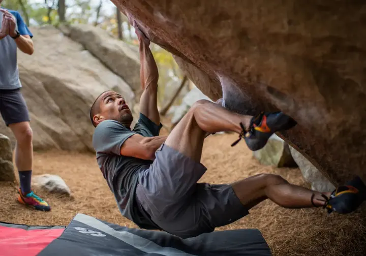 Boulderer demonstrating a safe falling technique (tuck and roll) as they land on a crash pad, highlighting the climber's active role in fall management and bouldering safety.