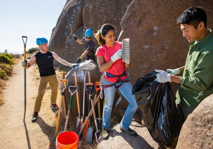Diverse group of climbers actively participating in crag care by doing trail maintenance and brushing holds, demonstrating practical protection of climbing environments.
