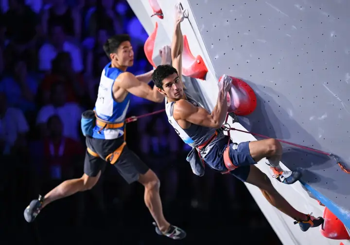 Two male speed climbers racing side-by-side up the standardized 15-meter wall in a vertical sprint during an IFSC competition.