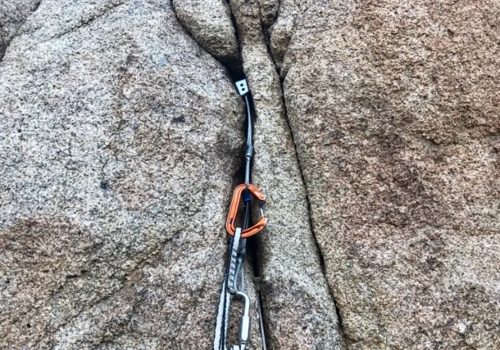 Close-up of a perfectly placed cam (SLCD) in a granite crack with a climber's hands, illustrating cams as workhorse active protection in a trad rack.