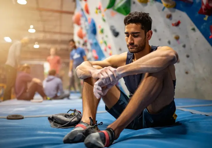 Climber sitting calmly at the base of a challenging indoor route, eyes focused or closed in visualization, cultivating a resilient climbing mindset for progressing skills.