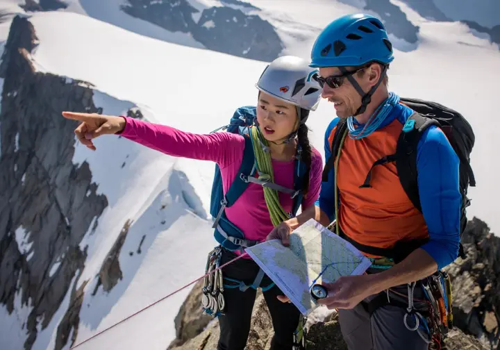 Two diverse alpinists using in-field alpine navigation techniques: one points to a route feature while the other consults a map and compass on a mountain slope.