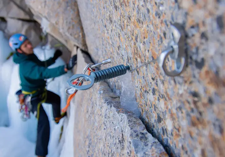 Close-up of mixed climbing protection: an ice screw securely in ice next to a cam or nut placed in an adjacent rock crack, with hands assessing the setup.