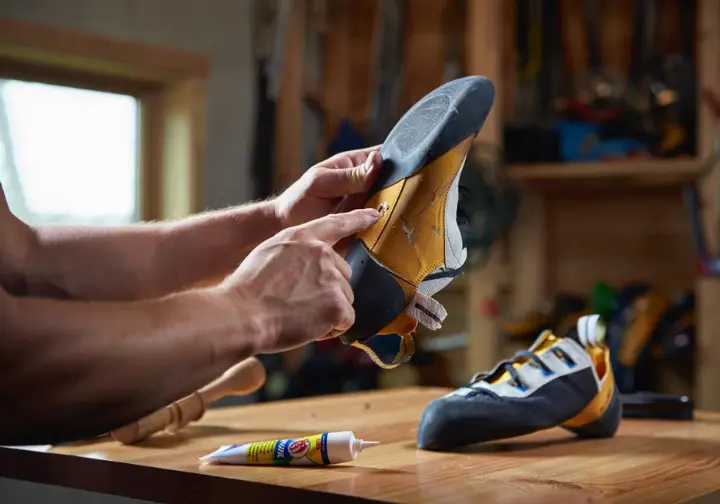 A climber inspects a worn climbing shoe toe, weighing the options to repair, resole, or replace it, with repair glue and a new shoe nearby.