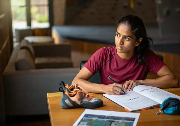Female climber at a table in a gym, structuring her indoor climbing training plan in a journal for effective results, surrounded by climbing gear.