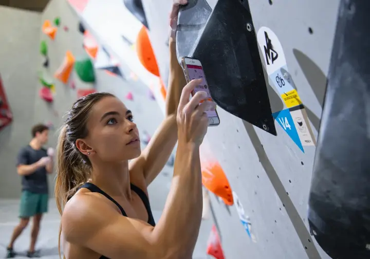 Climber in an indoor gym looking at a bouldering problem with a visible grade tag (e.g., V4 or 6B), understanding and gauging the challenge based on bouldering grades.