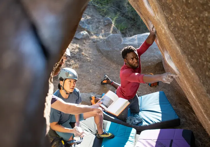 Two diverse boulderers at an outdoor crag conducting a holistic risk assessment: examining the boulder problem, discussing crash pad placement, and reviewing safety protocols before climbing.