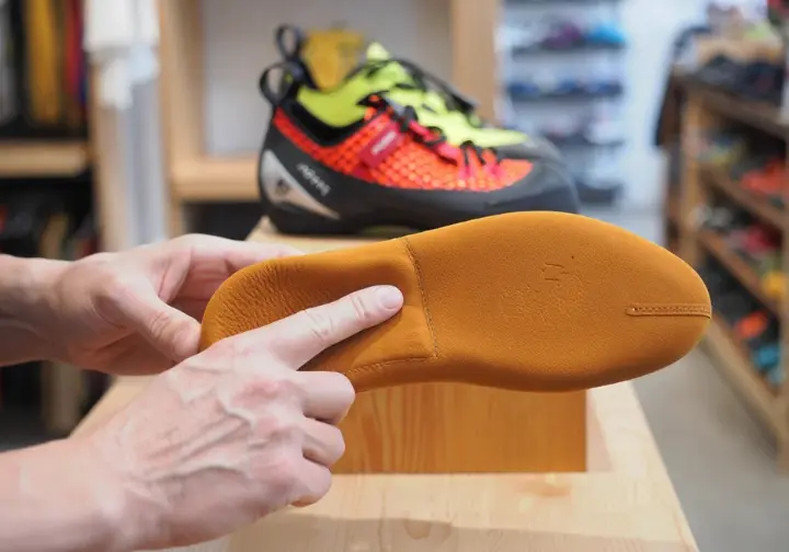 A climber holds a leather climbing shoe in the foreground with a synthetic model in the background, showing the material differences for shoe care.